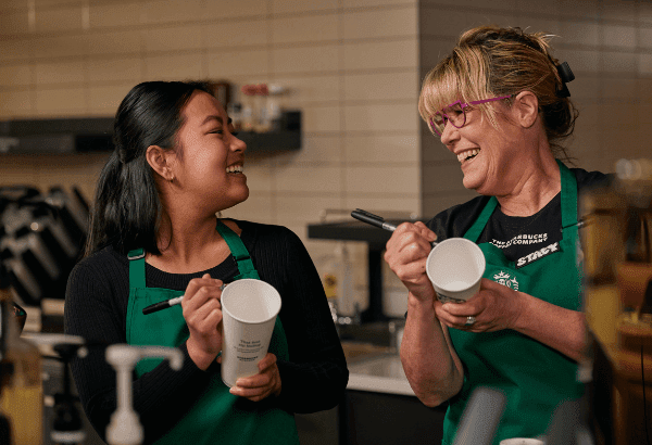 A Starbucks barista and her manager laugh together as they write on cups.