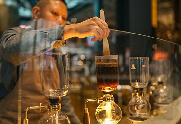 An individual standing at a coffee siphon, carefully stirring the contents with a long-handled spoon.