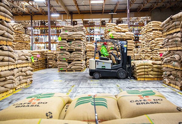 A person operating a forklift in a warehouse filled with neatly stacked burlap coffee bags, maneuvers through the warehouse