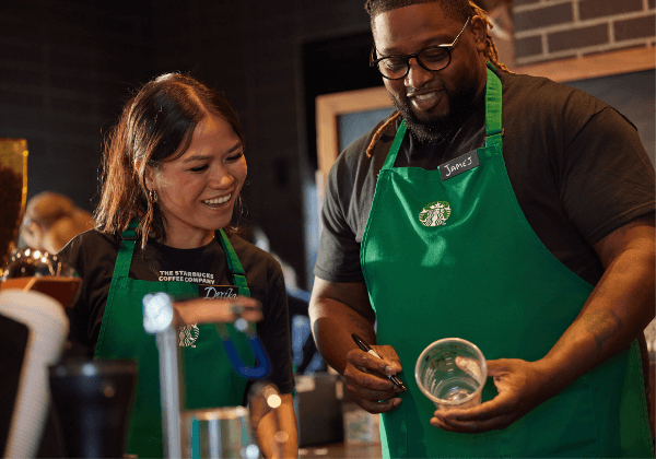 Two Starbucks baristas in a coffeehouse look at a cup that one has written on.