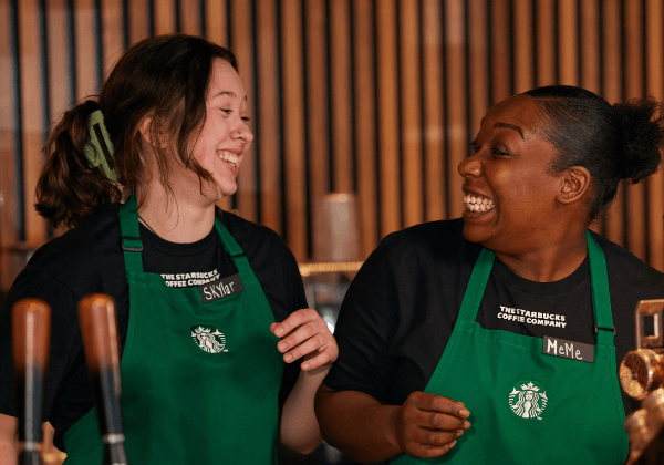 Two Starbucks baristas wearing green aprons look at each other and laugh in the moment.